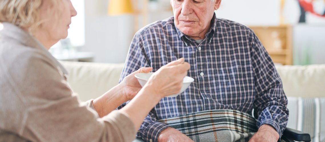 Senior grey-haired man in wheelchair looking at spoon with soup while his carer or wife feeding him by dinner